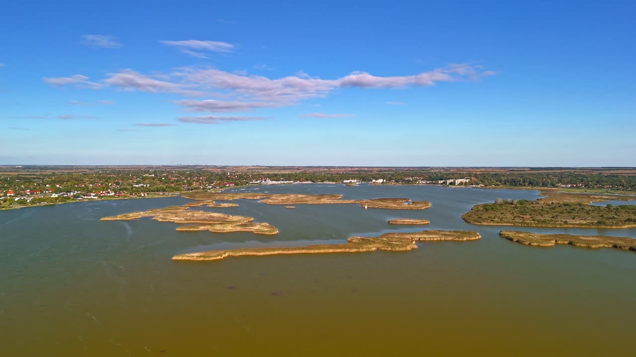 Drone hyperlapse above Lake Velence, Hungary, showing reed-covered islands, calm autumn waters and the lakeside towns under a vivid blue sky