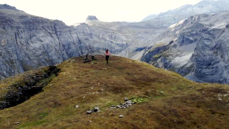 A-flyover-following-a-hiker-running-towards-the-view-of-lake-Limmernsee-in-Glarus,-Switzerland,-the-turquoise-colored-water-of-which-is-surrounded-by-tall-Swiss-Alps-peaks-and-cliffs