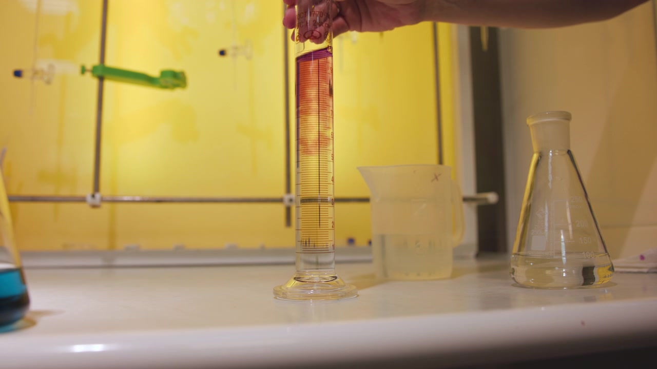 Mano guantata che regge un cilindro graduato mentre una goccia di reagente rosso si forma sopra un liquido trasparente all'interno di una cappa aspirante gialla