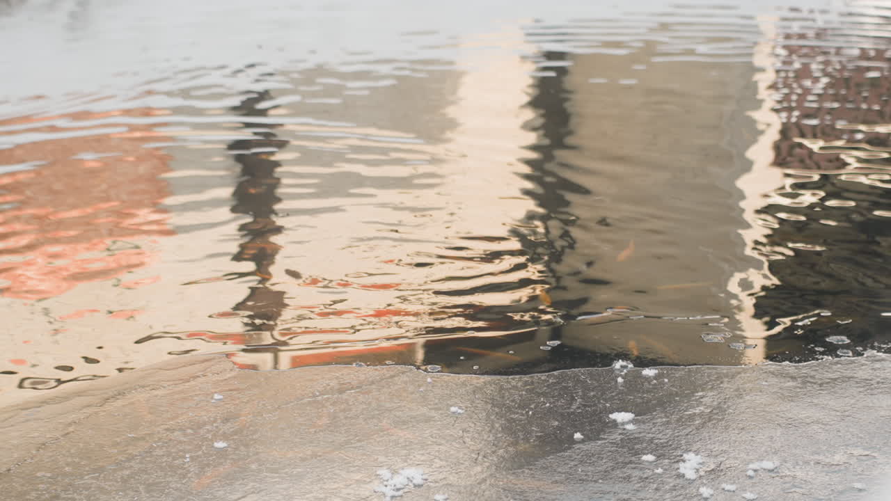 Puddle reflection of urban building facade, soft ripples across wet pavement reveal orange and beige window shapes, shallow water pooling on concrete sidewalk, subtle shimmer and distortion as breeze