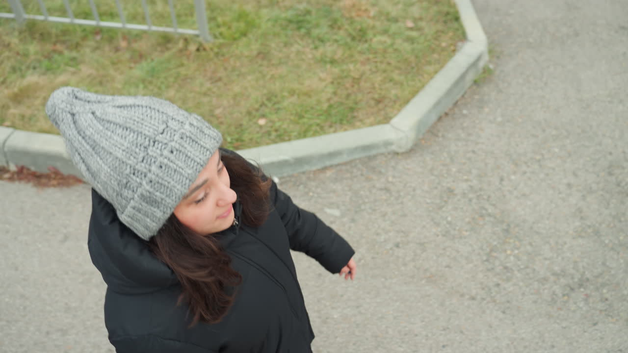 Donna urbana in abbigliamento invernale. Donna con berretto che balla gioiosamente sul marciapiede cittadino in inverno. Donna giocosa con berretto che si gode un ballo allegro su una strada cittadina nuvolosa in un paesaggio invernale