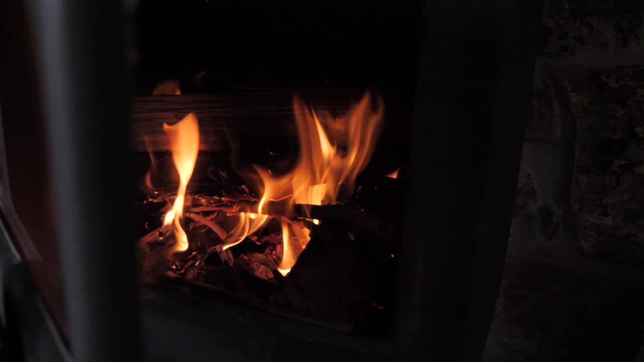 Red hot orange fire flame burns sticks and wood in small metal cabin fireplace in slow motion with sparks and embers flying up into flume.