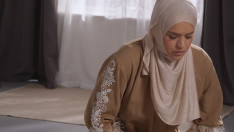 muslim woman wearing hijab at home praying kneeling on prayer mat 1