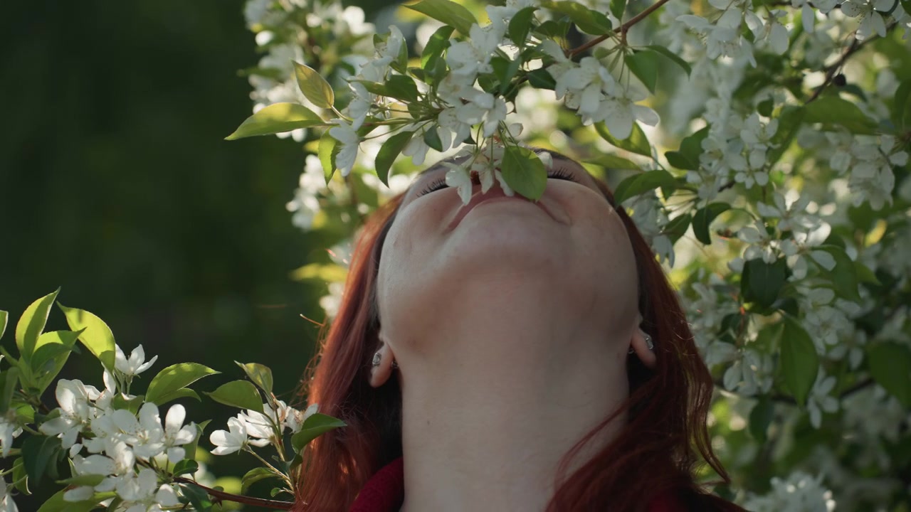 Primo piano di una donna con lunghi capelli rossi che indossa orecchini e annusa fiori bianchi su rami verdi. La scena presenta una luce naturale soffusa che si concentra unicamente sull'interazione tra il viso del soggetto.