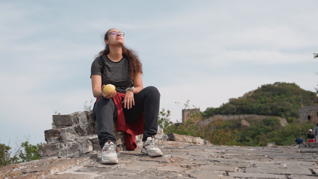 Giovane donna che riposa su antiche rovine, seduta su una pietra con zaino e bottiglia gialla, sneakers sulle macerie, fortezza distante e alberi sullo sfondo, postura rilassata e pausa di viaggio introspettiva