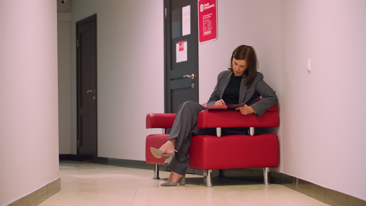 Businesswoman sitting on red chair in office hallway, deep in thought while reading and writing in notebook, wearing gray suit and heels, focused on task, calm professional office setting