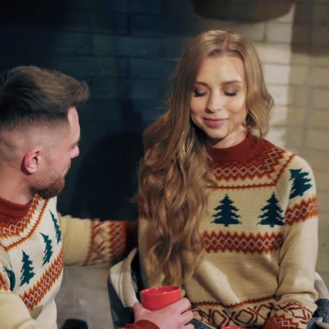 couple in love drinking tea. young man and beautiful girl in knitted christmas sweaters sitting near the fireplace. man kisses his girlfriend. romantic evening.