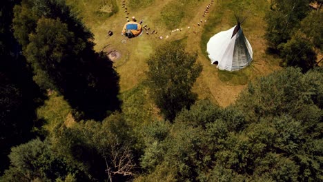 drone flies over a beautiful dense green forest to reveal a large outdoor event space with giant teepee, native american community