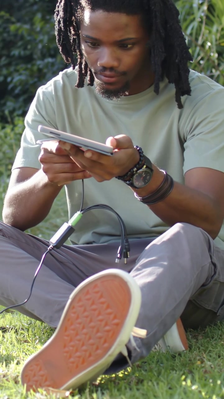Vertical video: Reaching, man in green tee plugging green plug into tablet on grass, checking link