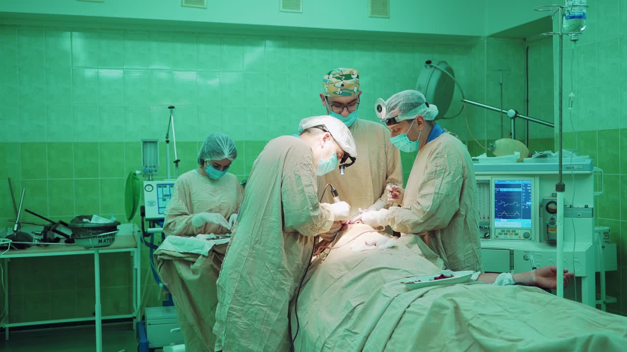 Teamwork of professional surgeons in the operating room. Doctors in special uniform work with medical equipment during the operation on a patient on the green background in clinic.