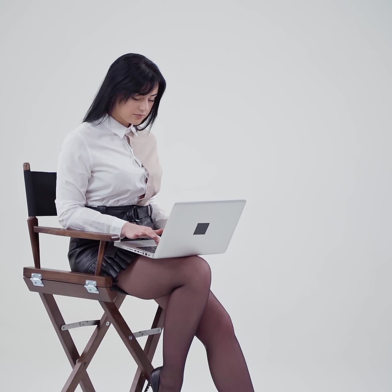 Sexy young lady open laptop in studio. Beautiful woman in white blouse and short skirt sitting on a chair and holding laptop on light background. Slow motion.