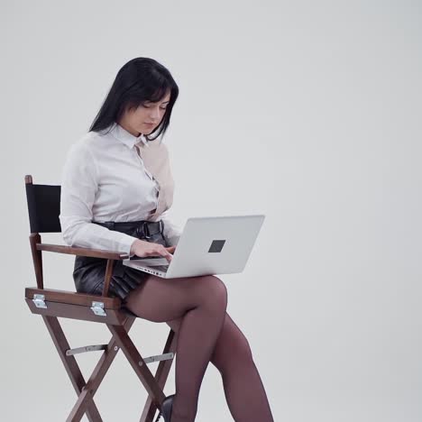 sexy young lady open laptop in studio. beautiful woman in white blouse and short skirt sitting on a chair and holding laptop on light background. slow motion.