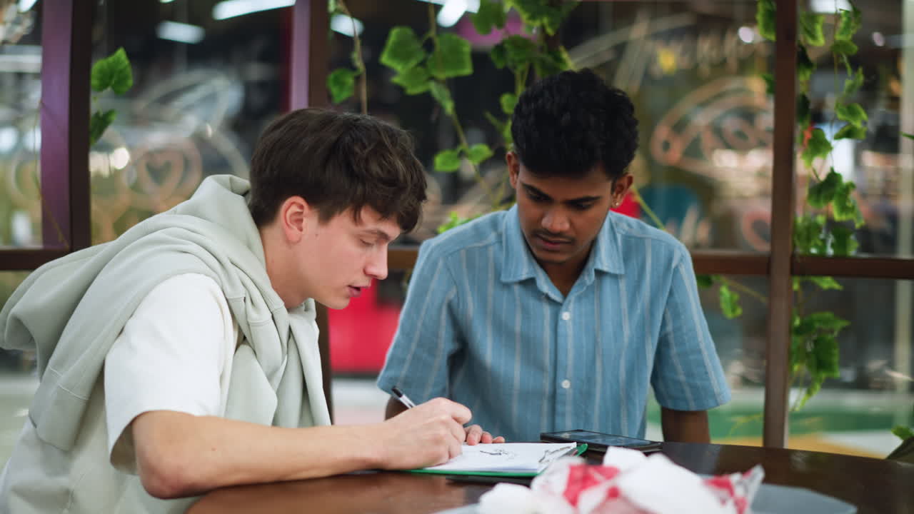 White boy leaning over sketch paper glancing at friend phone for drawing reference while shading pencil strokes in art session showing creative learning and focused attention on detail and form