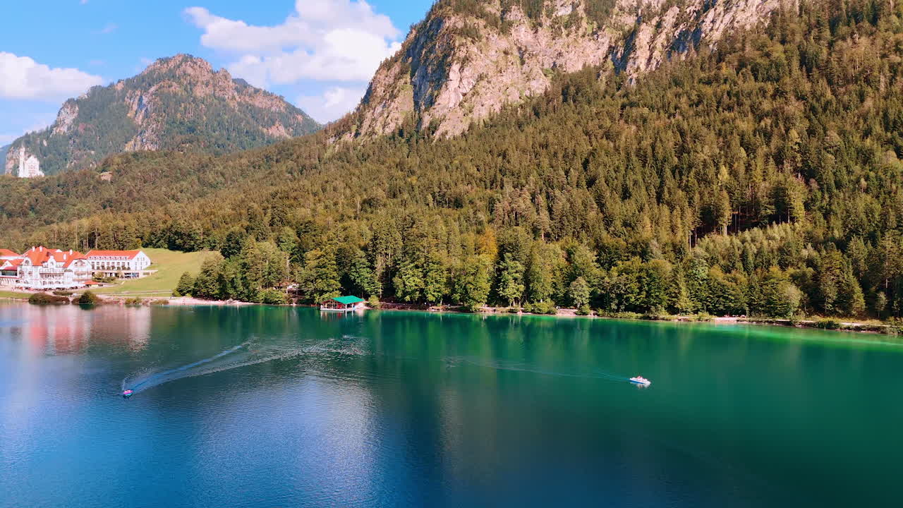 Turquoise lake with a boat. Pine tree woods grow on the rocks at the lakefront. Beautiful buildings and Neuschwanstein Castle at backdrop. Bavaria, Germany