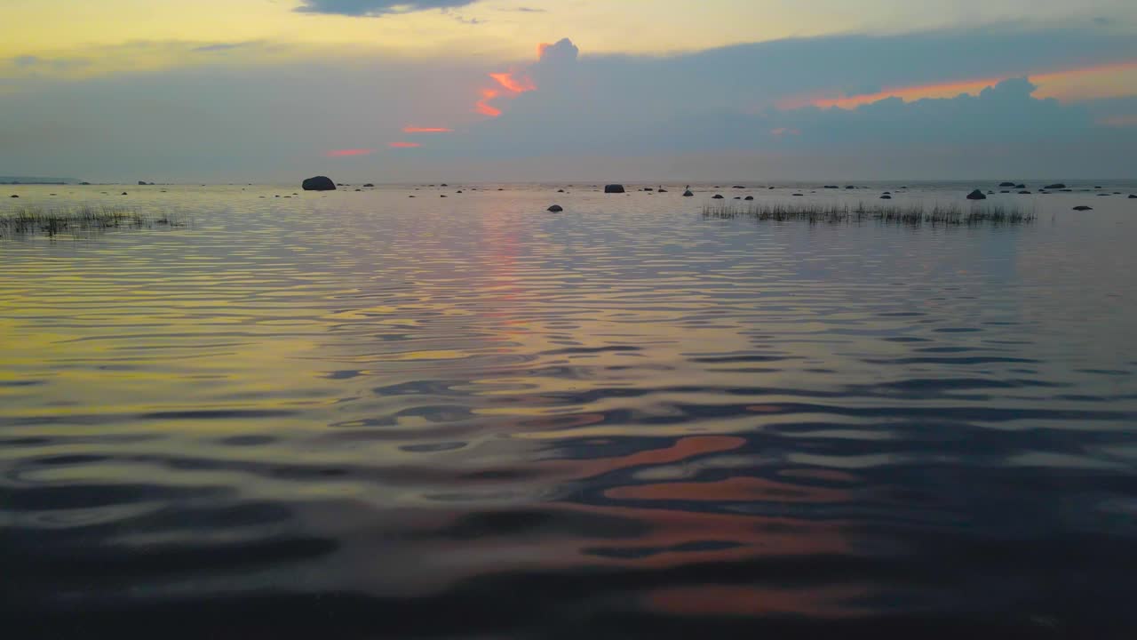 Aerial drone gliding and flying above ocean shoreline beach with reed and beach grass during a orange, red and yellow colored sunset in the evening. Rocks, boulders and rippling sea water waves seen
