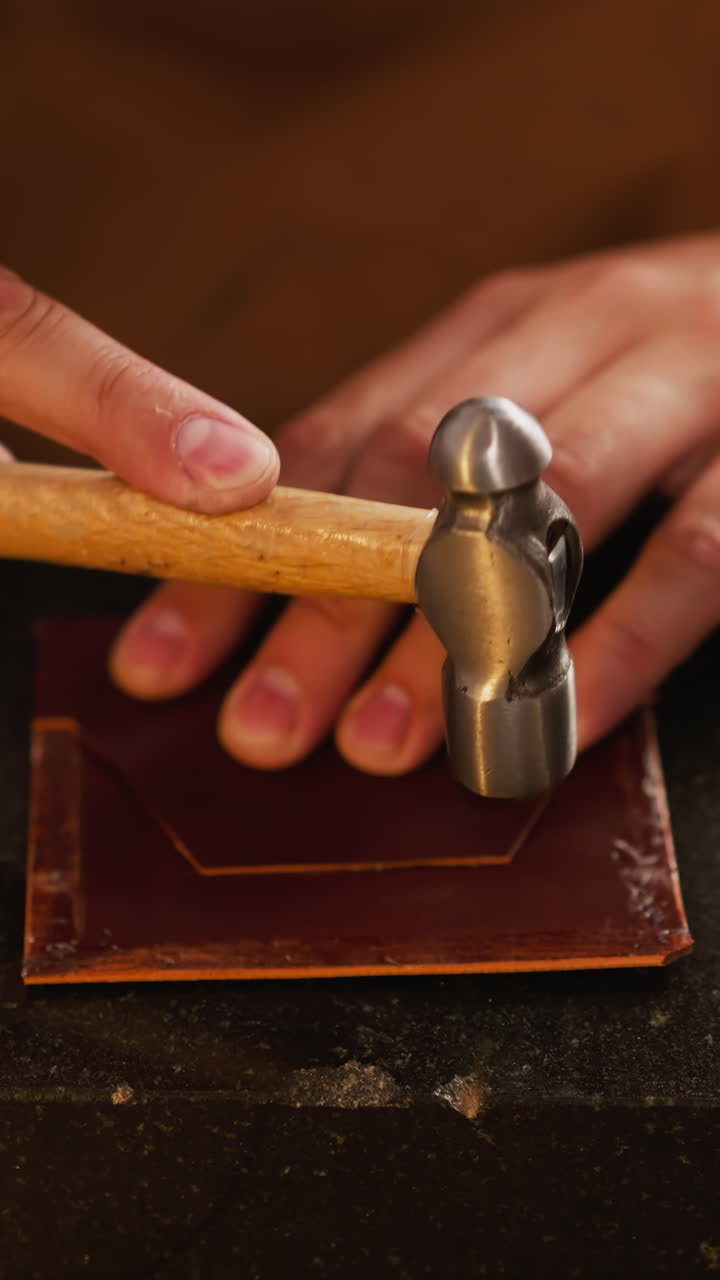 Skilled craftsman taps leather work-piece with hammer at table in workshop closeup. Traditional manufacture of wallets. Stylish handmade accessory