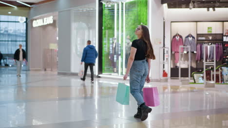 young woman with shopping bags in mall flaunts her hair and looks back confidently while other shoppers walk by in background, showcasing vibrant retail atmosphere and shopping experience