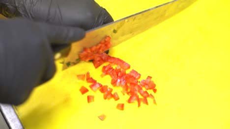 chef, wearing black sterile gloves, carefully and skillfully cutting, slicing, and dicing tomatoes in small cubes with a sharp chef's knife on a yellow plastic cutting board