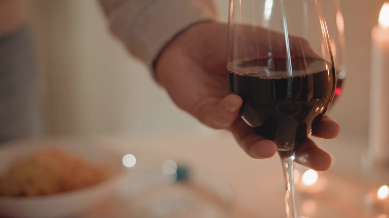 Secuencia en primer plano que muestra a una persona vertiendo líquido rojo oscuro de una botella verde en una copa de vino transparente, luego sosteniendo la copa llena por el tallo mientras un suave balanceo y el líquido ascendente crean ondas; ambiente interior cálido y suave.