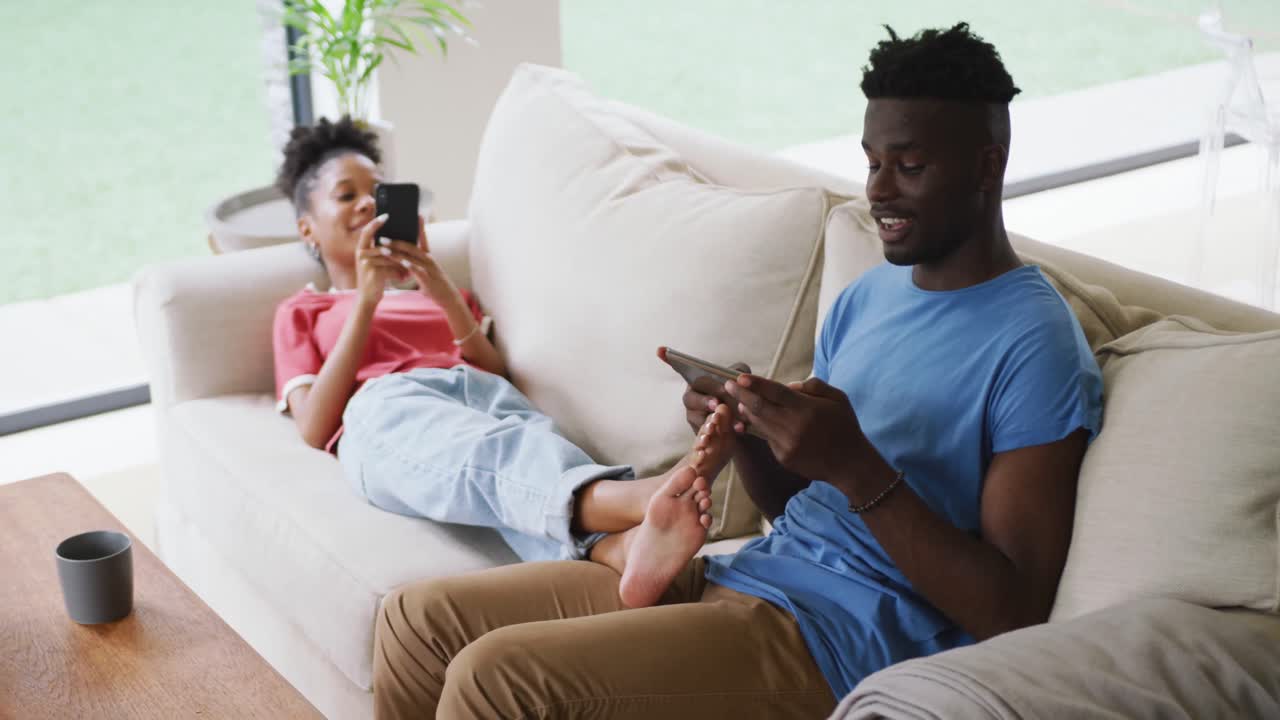 Video of happy african american couple relaxing on sofa with tablet and smartphone