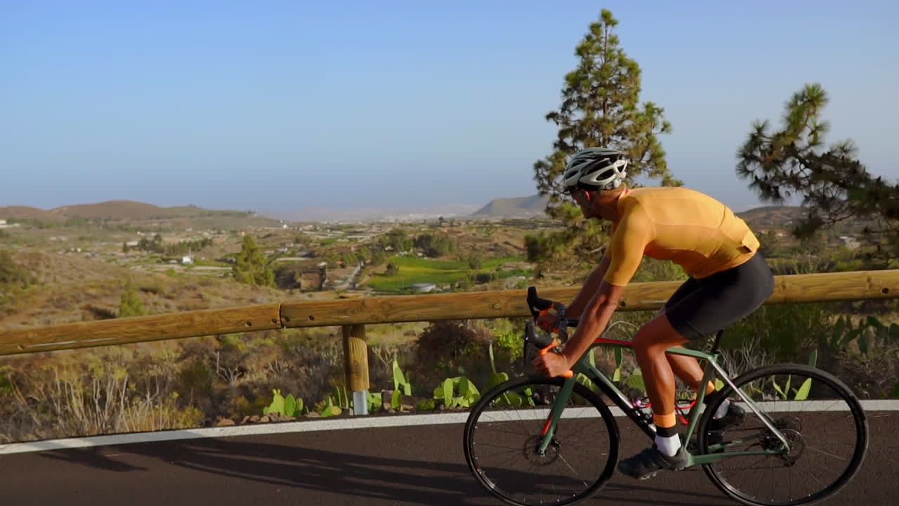 This tracking video showcases a male cyclist's ascent up a mountain road. His cycling training occurs on a sunny day along a hilly highway road