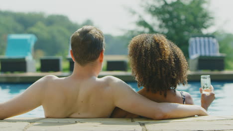 rear view of loving couple relaxing in swimming pool on vacation celebrating drinking champagne