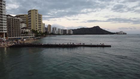 4k-Filmische-Drohnenaufnahme-Mit-Zoom-Von-Menschen,-Die-Den-Sonnenuntergang-Auf-Den-Felsen-In-Der-Nähe-Des-Waikiki-Strandes-Mit-Diamantkopf-Im-Hintergrund-Beobachten