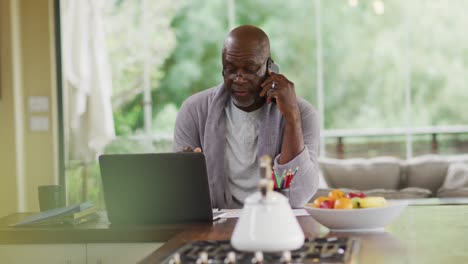 Afroamerikanischer-älterer-Mann-Im-Bademantel,-Der-In-Der-Küche-Sitzt,-Einen-Laptop-Benutzt-Und-Mit-Dem-Smartphone-Spricht
