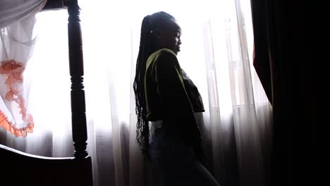 african woman putting on cropped belly shirt in front of a window of a hotel room - against light