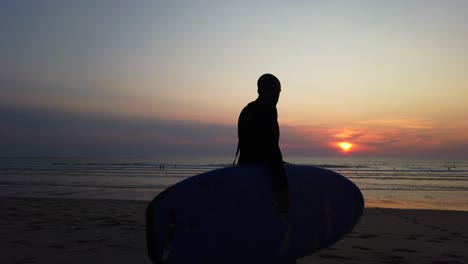 Silueta-De-Hombre-Con-Tabla-De-Surf,-Caminando-En-La-Playa-Contra-La-Hermosa-Luz-Del-Sol