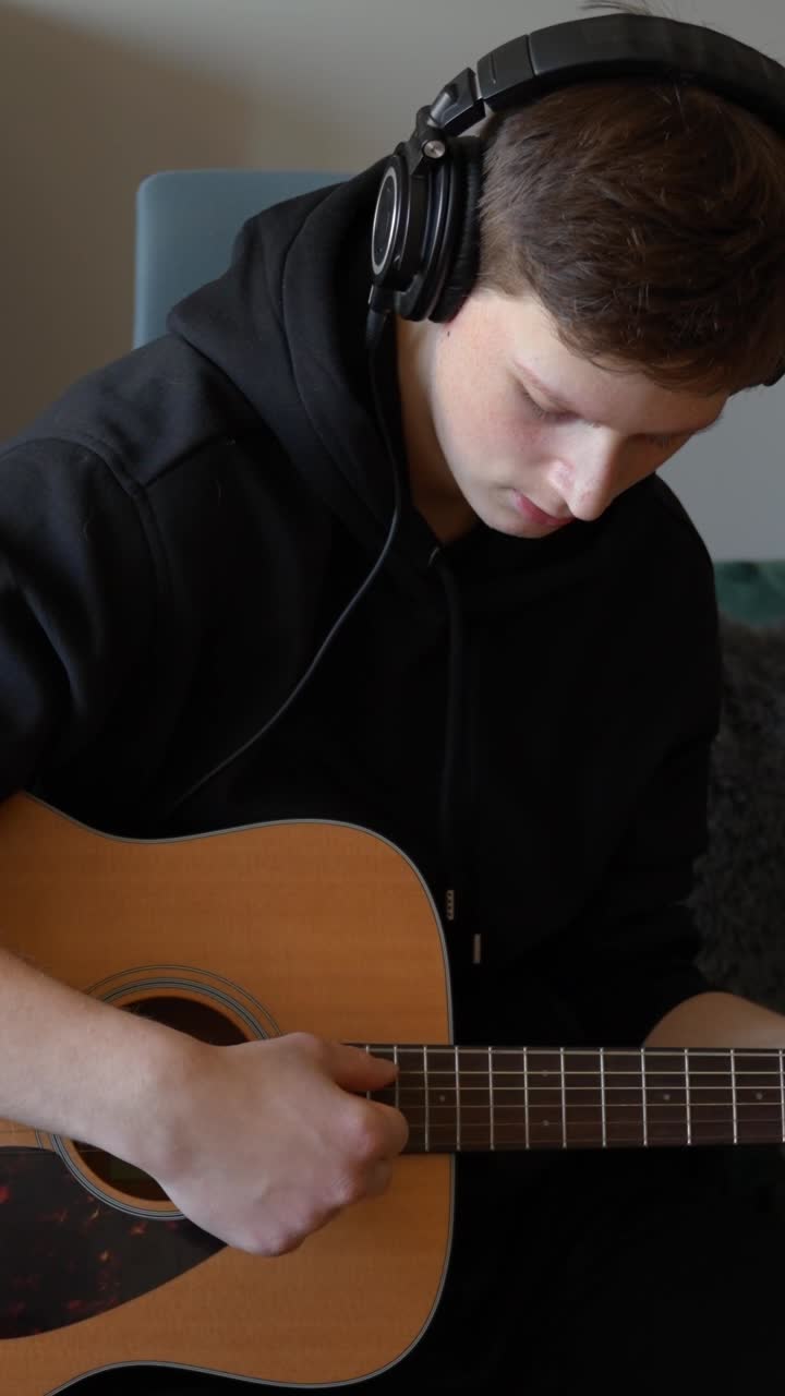 Young man wearing headphones and a black hoodie sitting, strumming an acoustic guitar, focusing on the instrument and creating music, practicing his hobby at home, vertical shot