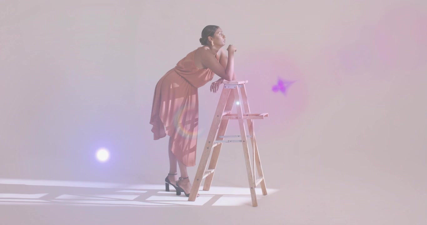 Donna che entra in un set di moda, cammina su una zona illuminata a griglia, si appoggia a una scala e contempla sfere vorticose.