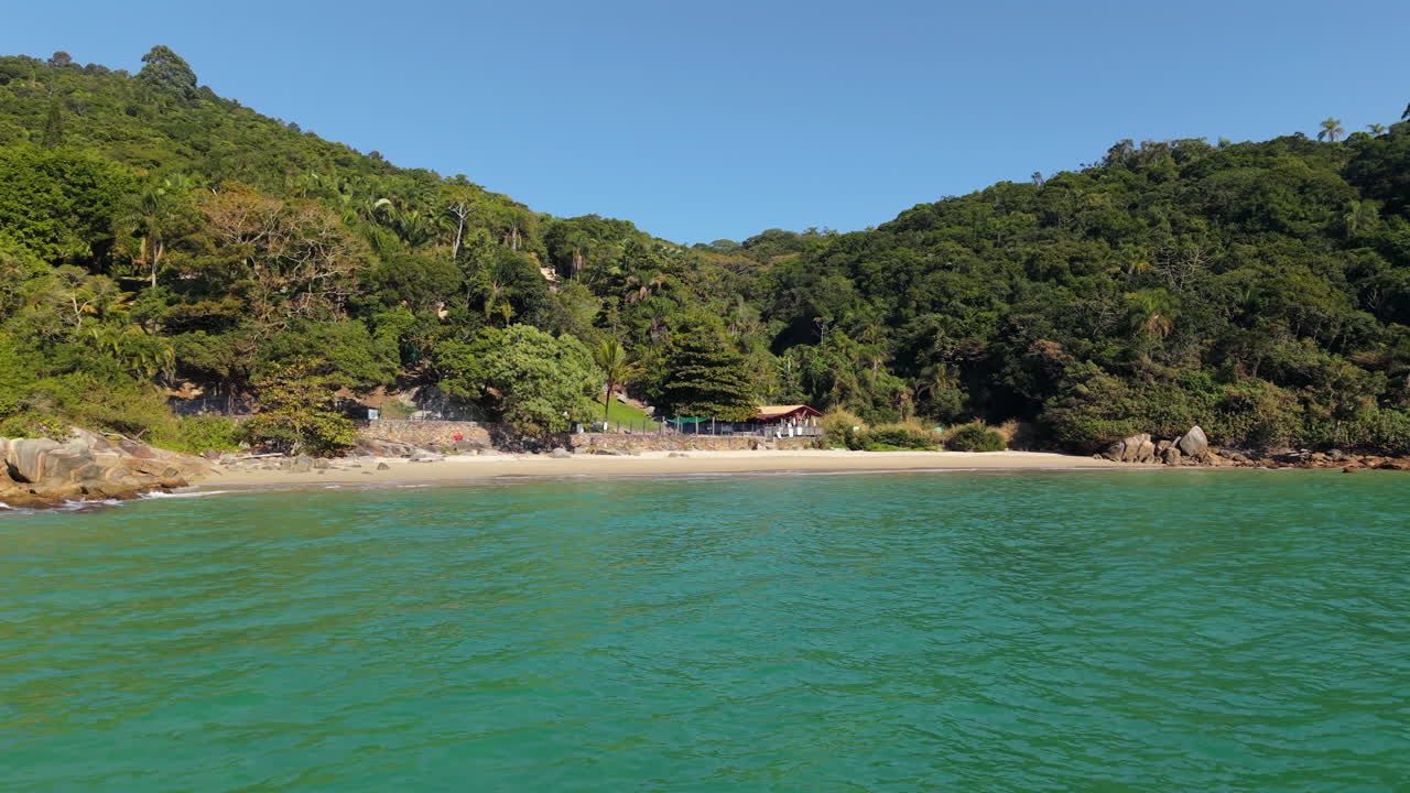 A stunning low aerial drone flight over clear emerald water towards the secluded Praia do Estaleiro beach in Santa Catarina, Brazil. A hidden tropical paradise
