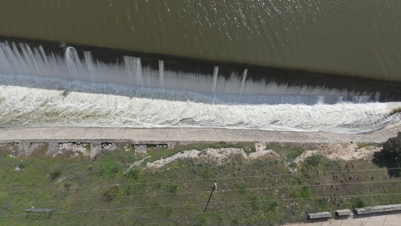 Lateral overhead drone shot showing a dam spillway releasing heavy water into a canal. Foam turns the flow white as it cascades beneath a bridge in a dynamic and powerful scene.