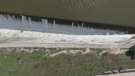 lateral overhead drone shot showing a dam spillway releasing heavy water into a canal. foam turns the flow white as it cascades beneath a bridge in a dynamic and powerful scene.
