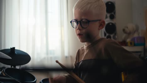 fotografía de cerca de un niño albino rubio con gafas tocando una batería electrónica usando palos especiales y escuchando música en su habitación. niño feliz persiguiendo su pasatiempo y desarrollando su talento para tocar instrumentos musicales en casa en su habitación