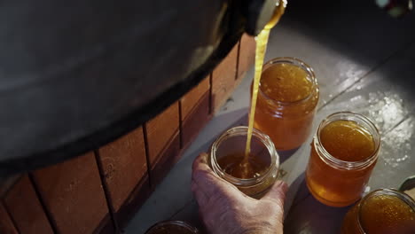 traditional honey production and harvest in bee farm, freshly extracted golden pure liquid honey flowing from extractor tank into glass jars, hand holding the jar