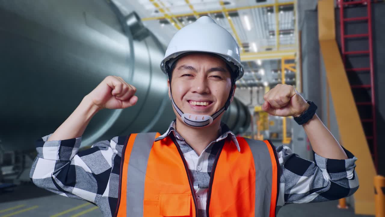 안전 헬을 착용한 아시아 남성 엔지니어가 파이프 제조 공장에서 혀서 카메라를 향해 미소 짓고 있다.