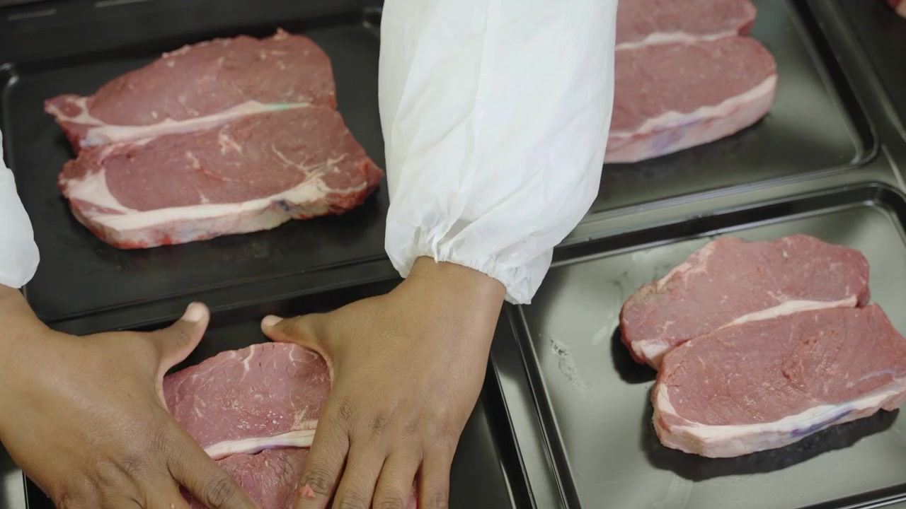 Uncooked beef steaks rest in individual sections of black plastic trays on a packing line in a modern meat facility.