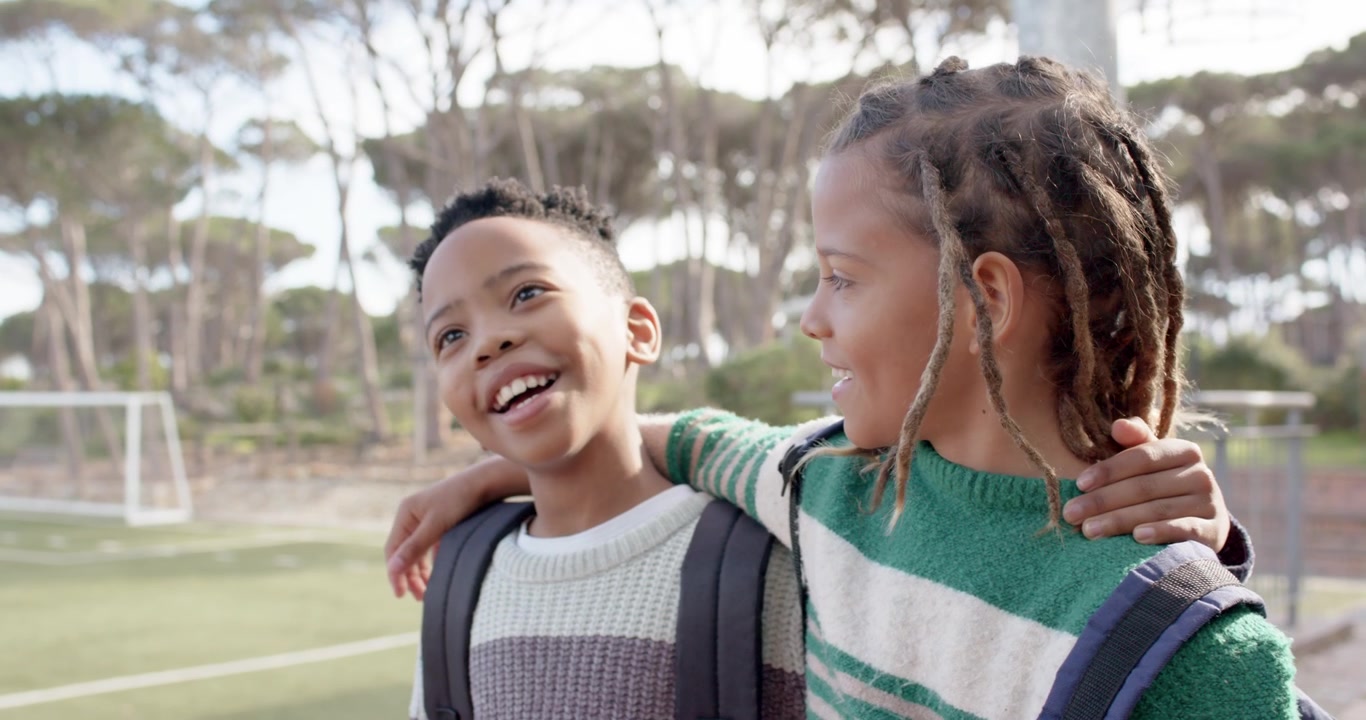 Meninos pré-adolescentes afro-americanos reagindo a um jogo de futebol em campo de grama sintética, conversando e sorrindo com mochilas.