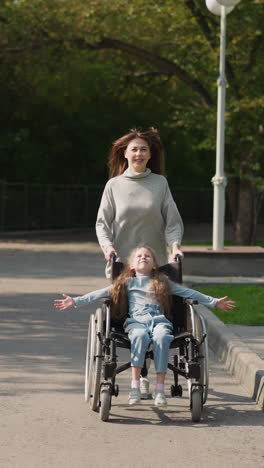 happy mother pushes daughter in wheelchair running on pavement in city park. woman and little girl with spinal paralysis enjoy sunny spring day