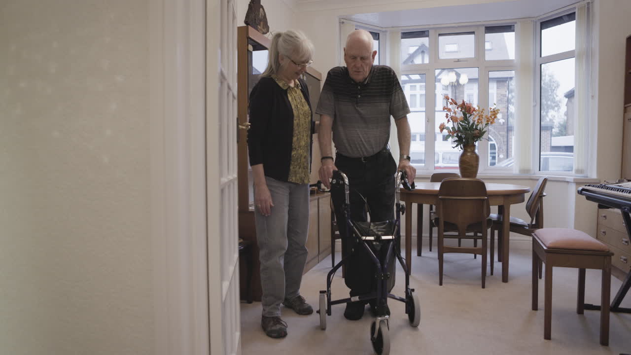 Elderly man using a walker with a woman assisting him at home