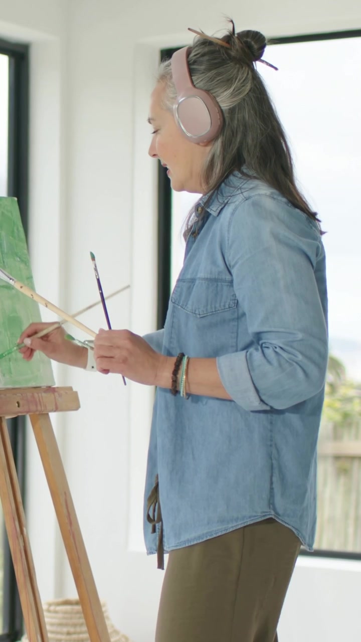 Video vertical: Mujer observando el lienzo, pintando en un caballete, terminando una obra de arte en casa con auriculares rosas.