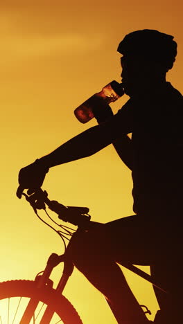 silhouette of professional bicyclist taking a bottle and drinking water at golden sunset. male cyclist in helmet drinks water on the natural background of setting the sun in the evening. vertical video