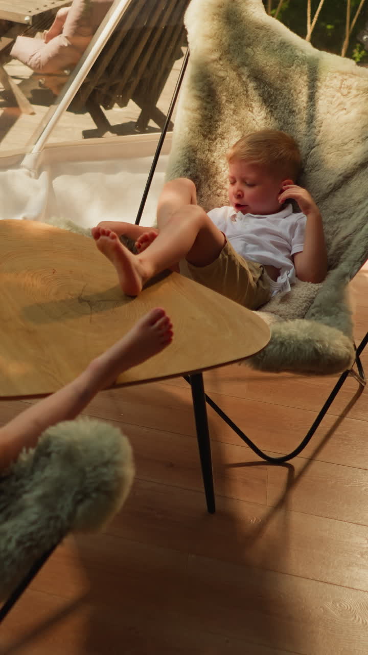 Children sit in furry chairs in glamping cabin. Yawning boy and sister enjoy lazy afternoon on shady hotel terrace. Little kids at low table