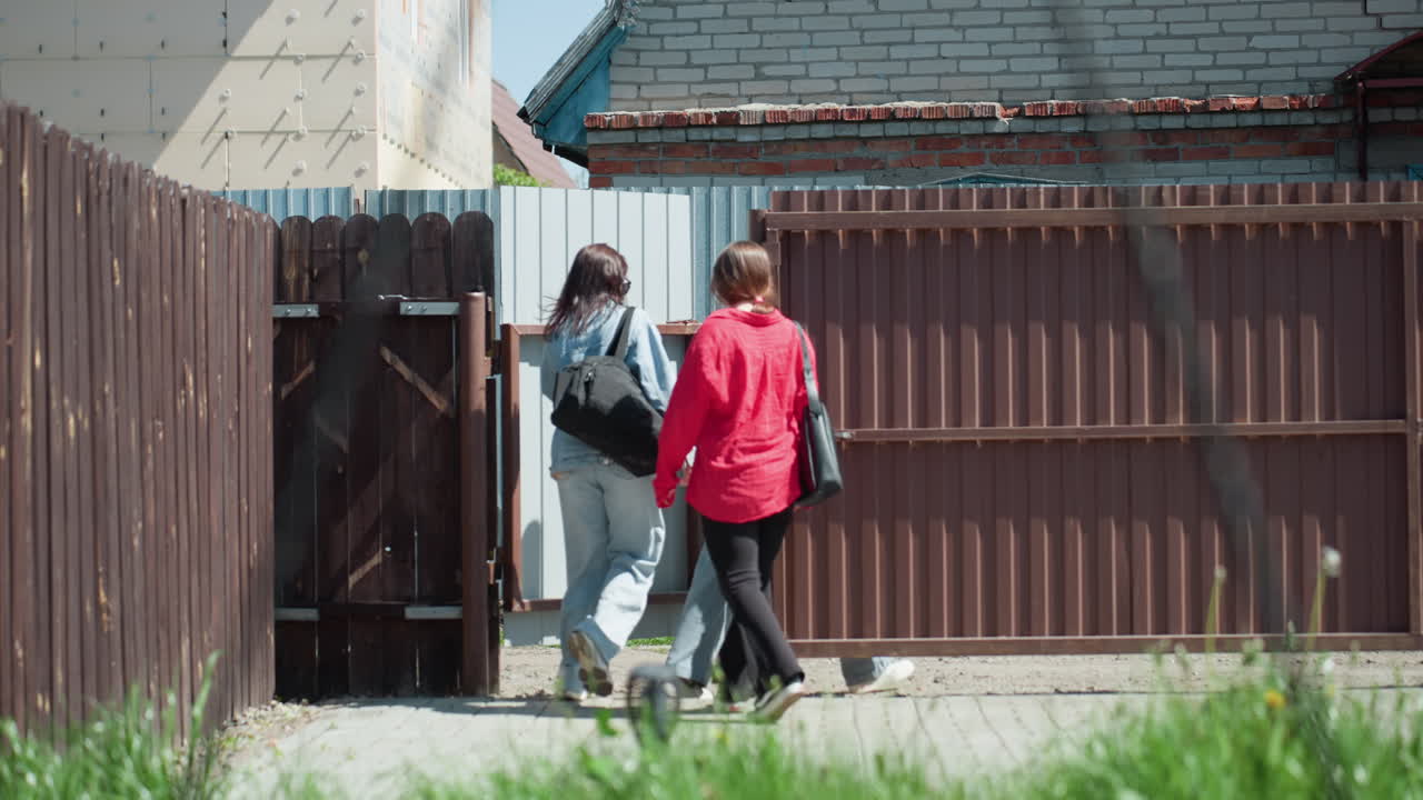 Group of young women walking into compound after shopping, one carrying paper bag, last person in red shirt seen closing gate, warm sunny day with fence, brick building, and grass in foreground