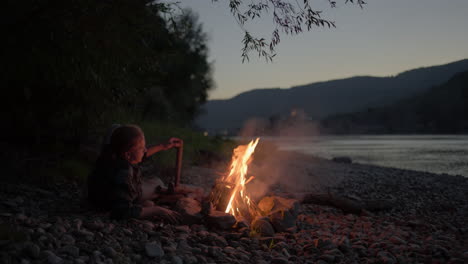 Man-lays-down-relaxed-next-to-bonfire-on-pebble-beach-by-the-river,-slow-motion-blue-hour