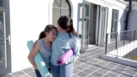 happy biracial mother and daughter standing with yoga mats on terrace in sunny day, slow motion