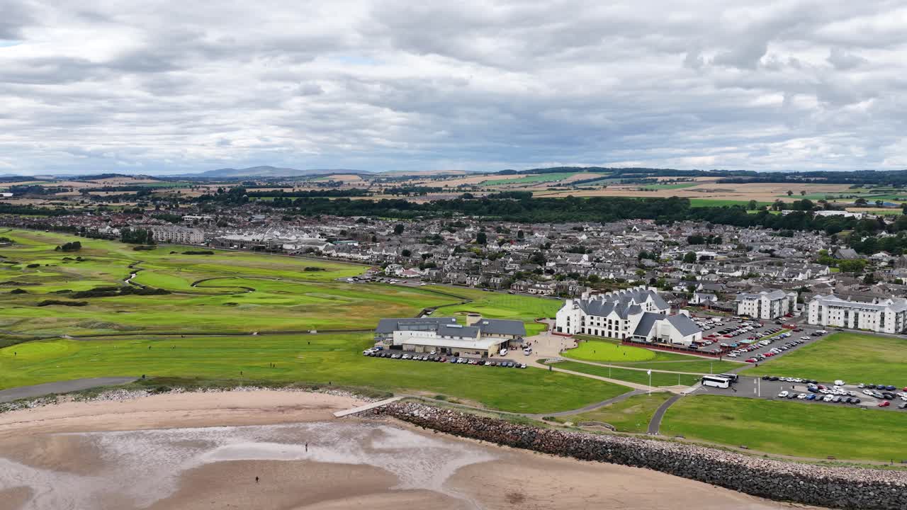 Drone pans over lush golf course, sandy beach, and town under overcast daylight, wide perspective