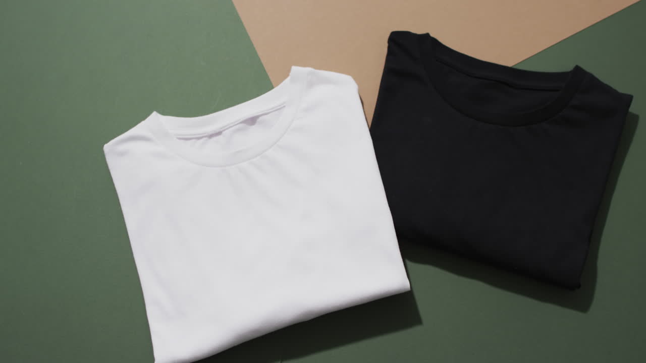 Video of flat lay of folded white and black t shirts with copy space on green and brown background